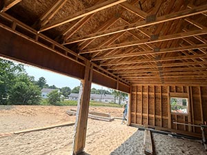 Open wooden-framed structure under construction with a sandy floor and exposed beams; trees and houses visible beyond.